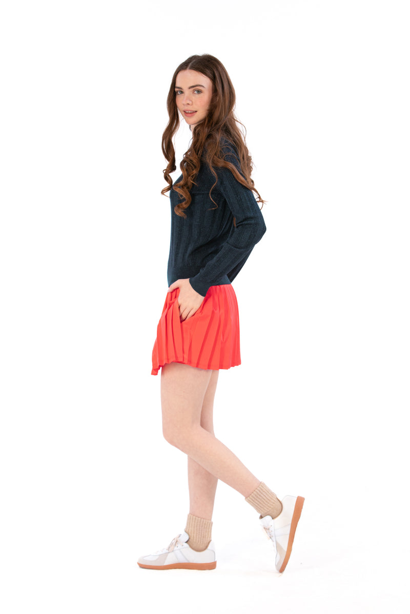 Woman wearing a navy sweater and coral pleated skirt on a white background