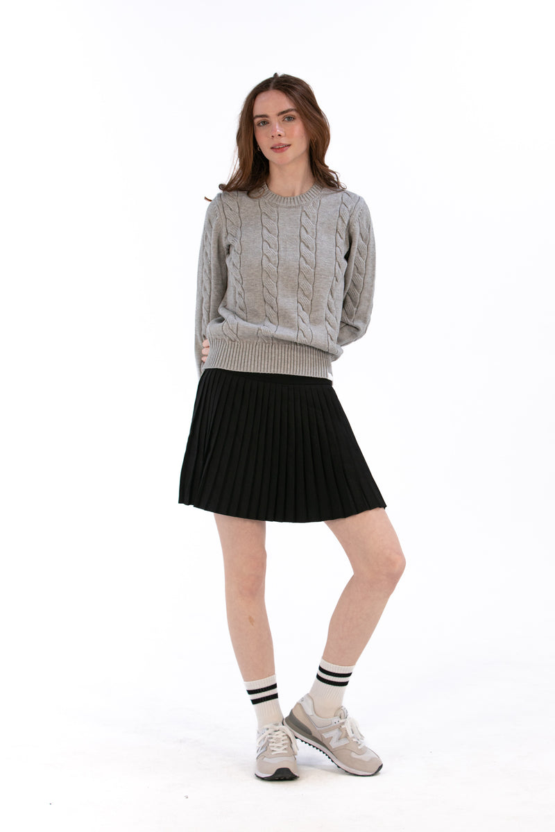 Woman wearing a marl gray cable knit sweater and black pleated skirt on a white background