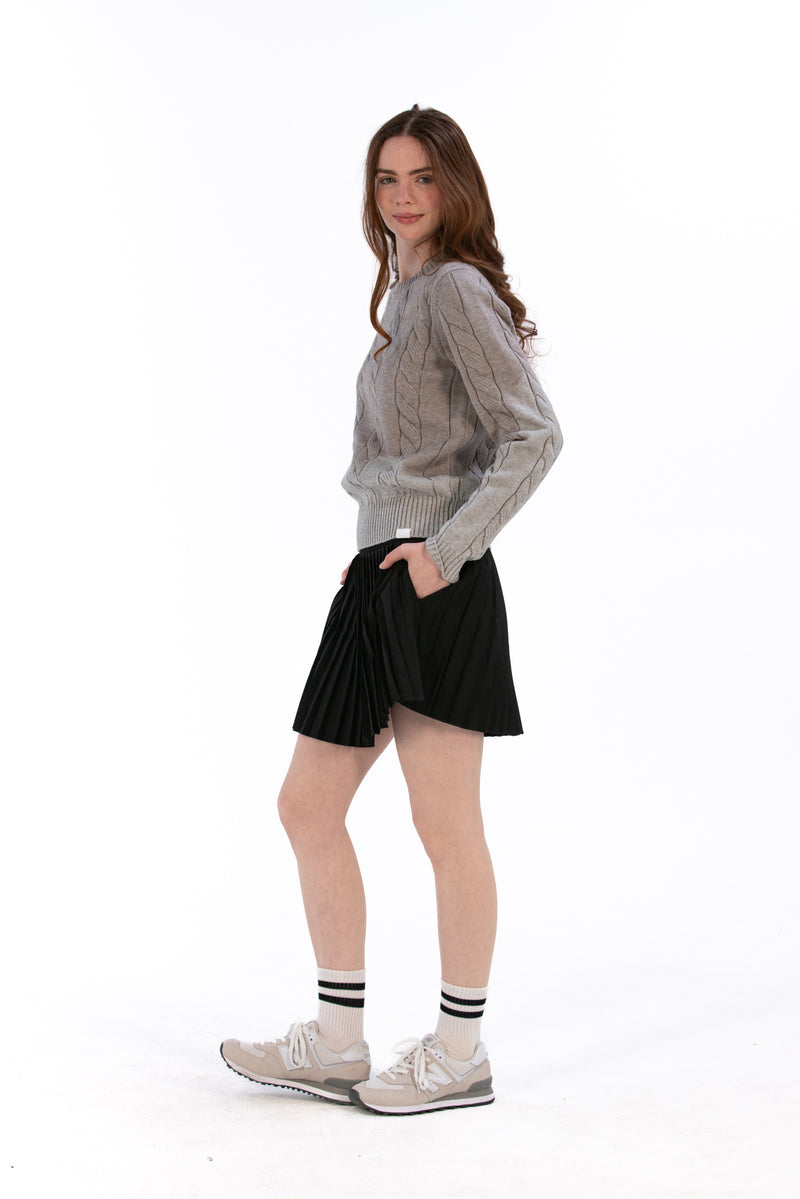 Woman wearing a marl gray cable knit sweater and black pleated skirt on a white background