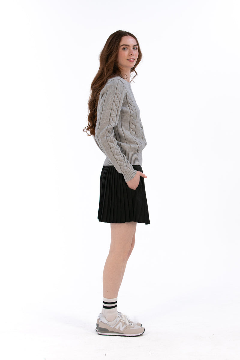 Woman wearing a marl gray cable knit sweater and black pleated skirt on a white background