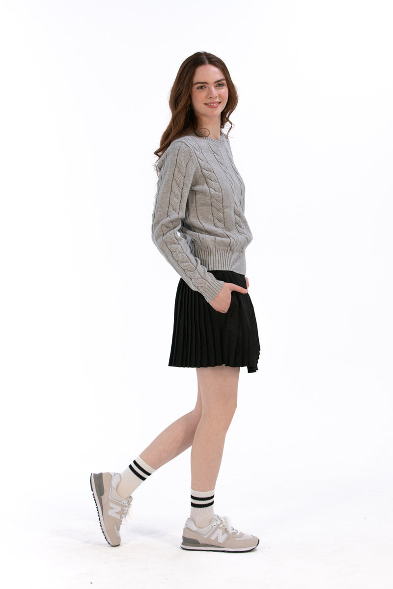 Woman wearing a marl gray cable knit sweater and black pleated skirt on a white background