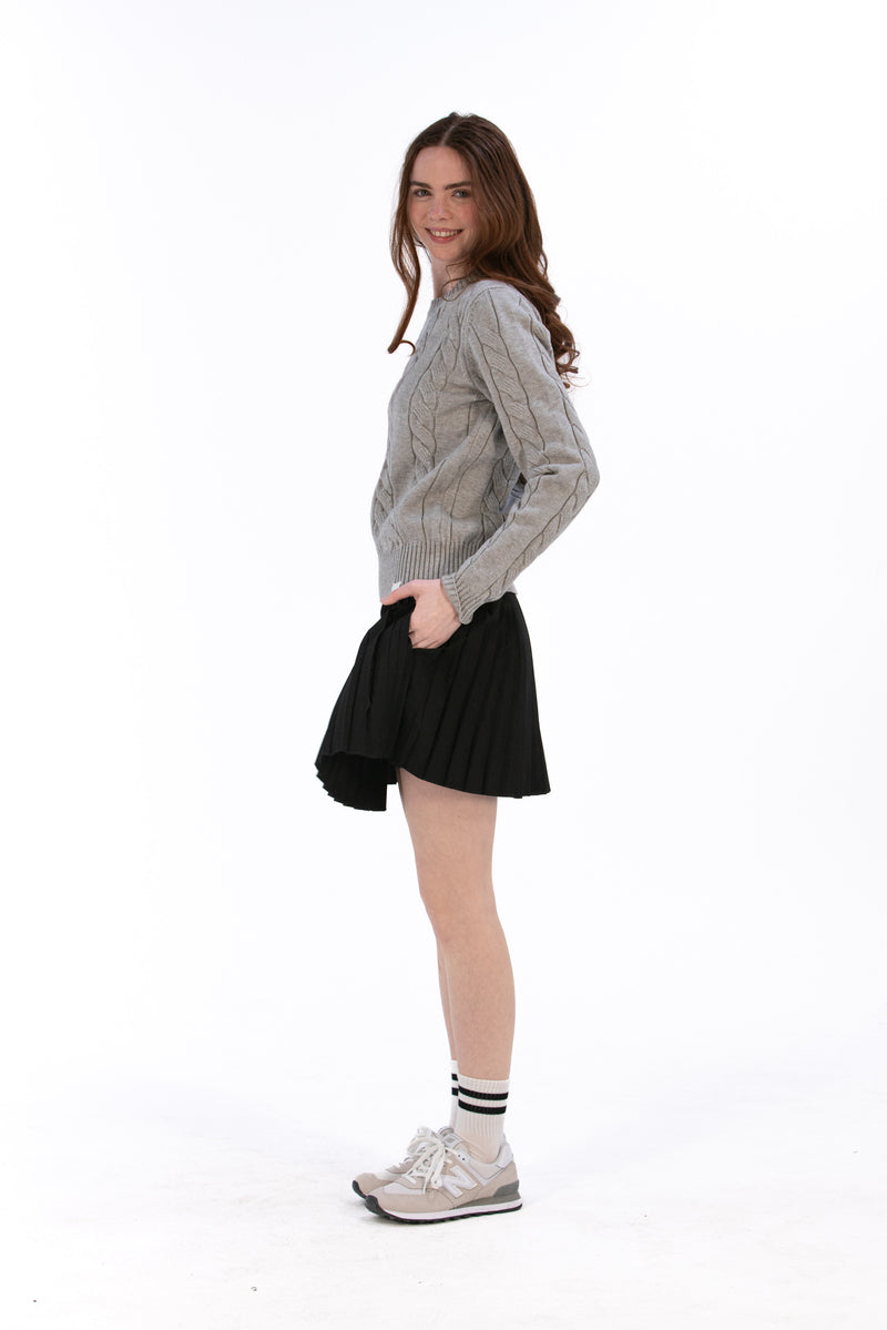 Woman wearing a marl gray cable knit sweater and black pleated skirt on a white background