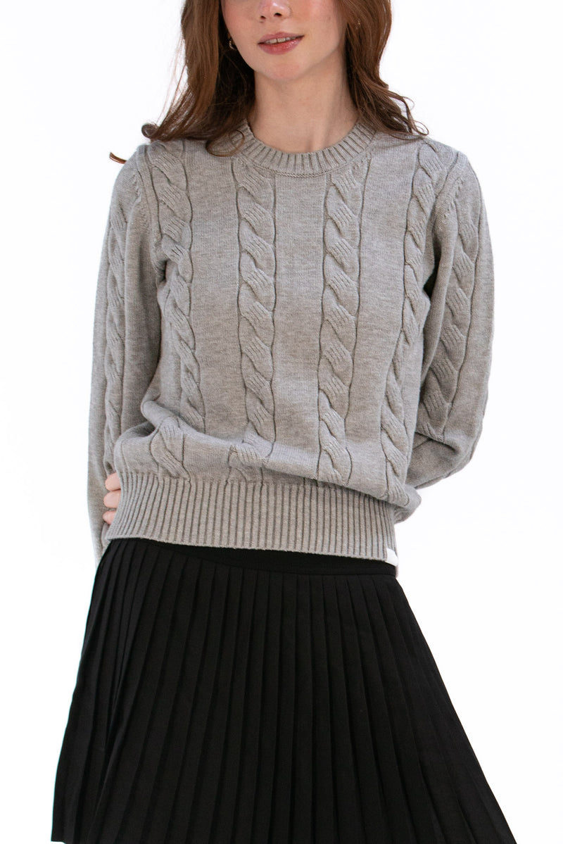 Woman wearing a marl gray cable knit sweater and black pleated skirt on a white background